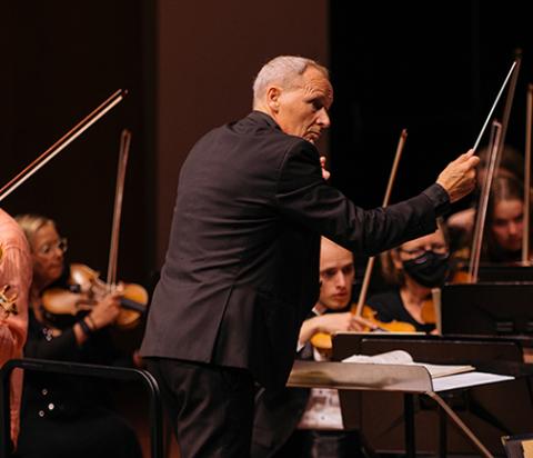 A conductor directs amid the violin section of the orchestra