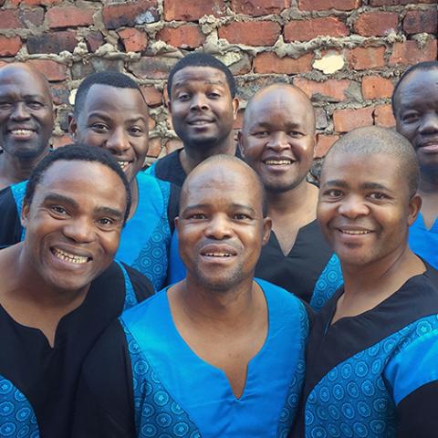 Eight members of Grammy Award-winning Ladysmith Black Mambazo, wearing blue, pose for a photo in front of a brick wall