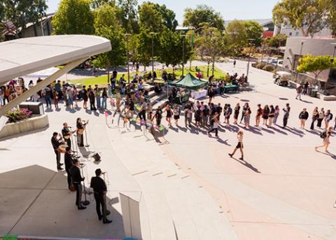 A line of students curves in a line in the University Union Plaza while performers play music on stage