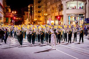 The Mustang Band, in white, green and gold uniforms, marches up a brightly-lit nighttime street in San Francisco