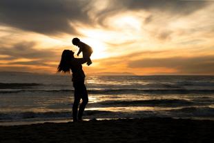 A woman holds a baby above her head on the beach at sunset.