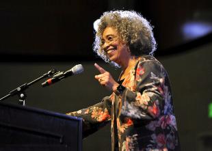 Activist Angela Davis holds up one finger and smiles while giving a talk at Cal Poly.