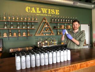 Alumnus Aaron Bergh holds up a bottle of his company's hand sanitizer, behind the bar at his distillery tasting room
