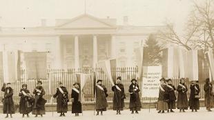 Photograph of fourteen suffragists in overcoats on picket line, holding suffrage banners in front of the White House. One banner reads: "Mr. President How Long Must Women Wait For Liberty". White House visible in background.