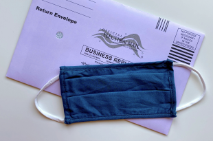 A picture of a mask on top of a vote-by-mail ballot.