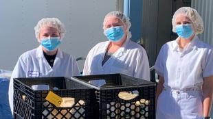 Three people wearing face masks and hair nets stand behind crates of food