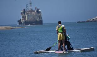 Cal Poly students paddle a raft on Morro Bay as an Army Corps of Engineers ship dredges the harbor in the background