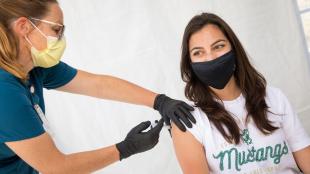 A student wearing a mask receives a vaccine from a health care worker wearing a mask and blue scrubs