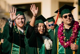Three students wear green graduation robes and hats and look at the camera during fall commencement.