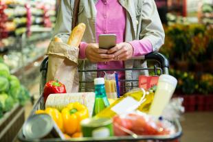 A woman texting on her phone at the grocery store. Her arms are resting on the handle of her shopping cart, which is full of food.