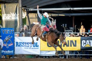 A man in a cowboy hat and a green vest rides a horse as it bucks in the air.