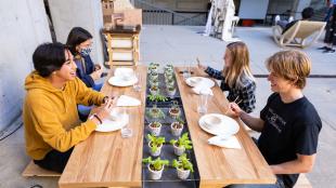Four students, including one wearing a mask, sit at a table with plates and green plants at an architecture exhibition