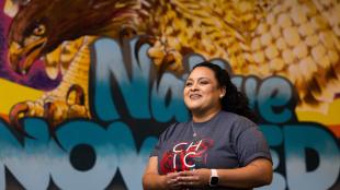 Cheryl Flores in front a colorful mural featuring a red tail hawk and the words 'Rooted in Native Knowledge'