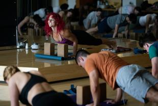 A yoga teacher in a purple tank top smiles as she poses in a plank position about to chaturanga in front of a yoga class at the rec center.