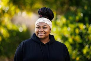 Nailah DuBose smiles for a portrait on Dexter Lawn. She is wearing a white headband and a black hoodie.