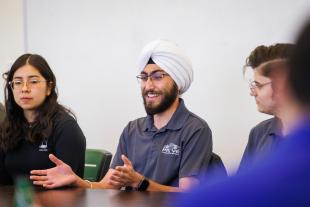 Three students sit at a table having a discussion. The middle student, who is wearing a turban, leads the discussion.