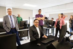 Congressman Salud Carbajal, center, sits at a desk with a computer in front of him while Cal Poly President Jeffrey Armstrong, Engineering Dean Amy Fleischer, Professor Paul Iscold and other people stand behind him. 