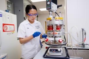 Grad student Ninjing Hua, in a white lab coat, latex gloves and safety glasses, loads a sample of olive pomace into an analysis device in a lab.