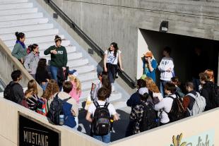 A group of people stand listening on a concrete balcony while a woman in a green Cal Poly sweatshirt speaks to them from the adjoining stairs.