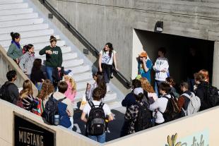 A group of people stands on a concrete balcony as a woman in a green Cal Poly sweatshirt speaks to them from the adjoining stairs.