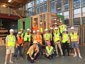 A group of students in safety vests and helmets pose in front of a partially-constructed tiny home