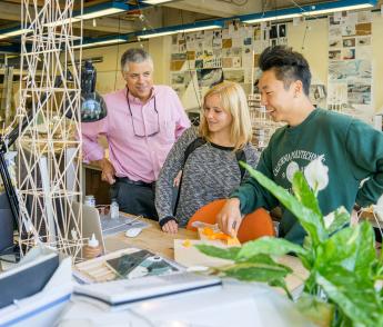 A man in a pink shirt, a woman in a grey shirt and a man in a green shirt inspect a structural model in a busy-looking architecture lab.
