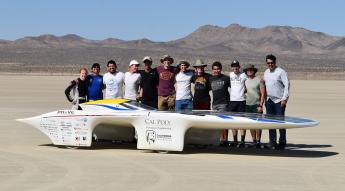 A group of students stands around a slim, futuristic-looking vehicle on a desert testing ground