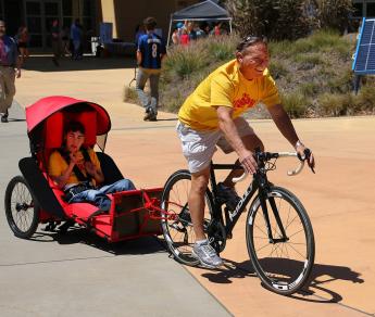 Joseph Cornelius rides in a bike trailer towed by his father, who is riding a bike. Both are wearing yellow shirts.