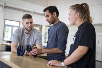 Three biomedical engineering students gather around a table to look at a prototype of a prosthetic thumb.