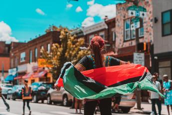 A woman in a dashiki-style shirt and long braids walks down a sunny city street in a parade, waving a red, black and green flag behind her