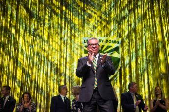 President Armstrong, onstage at a donor event in front of a green and gold backdrop, claps with a group of university supports behind him