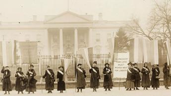 Photograph of fourteen suffragists in overcoats on picket line, holding suffrage banners in front of the White House. One banner reads: "Mr. President How Long Must Women Wait For Liberty". White House visible in background.