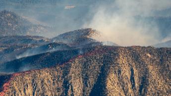 Smoke can be seen rising from behind a brown mountain, which has a red line of phos-chek along the top.