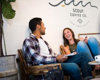 A man with short black hair and a plaid button down sits in a chair, while a girl wearing jeans sits in another chair to his right. Behind them is a white wall with Scout Coffee Co. painted on it under a drawing of ocean waves.