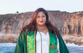 A woman in a white dress, red lipstick and reddish dark hair smiles and wears a green Cal Poly graduation robe and green and gold Cal Poly graduation stole. 