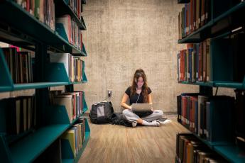 A woman in sweatpants and a black shirt looks at a computer with earphones in, studying, and sits against a wall between stacks of books in the Kennedy Library, with a big backpack next to her.