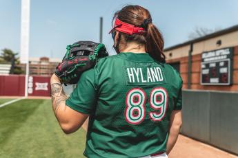 Sierra Hyland, a softball player for Mexico, faces away from the camera. She has a catching mitt on one hand and is wearing a mask. Her green jersey says Hyland and the number 89.