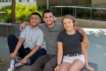 Three Cal Poly students sit on a bench outside. The male student in the center has his arms around another male student, left, and a female student, right.