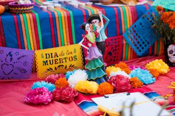 A figurine of festively-dressed skeletons dancing stands next to a cutout flag reading "Dia de los Muertos," surrounded by colorful cloth drapes and bright multicolored marigolds