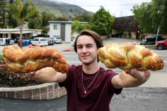 A man in a maroon shirt holds his arms outstretched, a challah loaf in each hand.