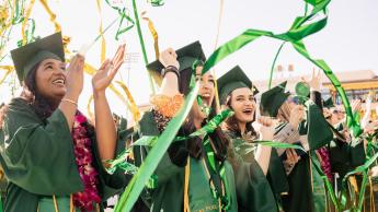 Graduates in green caps and gowns cheer while streamers fly in the air