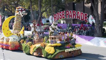 A Rose Parade float covered in flowers featuring cows with jetpacks drives down a sunny street with a Rose Parades sign behind it