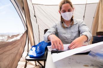 A woman wearing a facemask and sitting inside a tent holds something in a sock on top of what appears to be a binder.