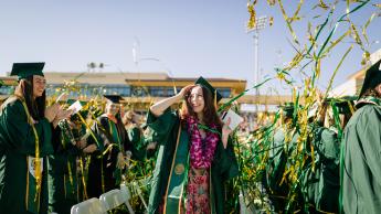 In a crowd of graduates at Spanos Stadium, a young woman in a green commencement gown and purple lei touches her mortarboard as green and gold confetti sail through the air
