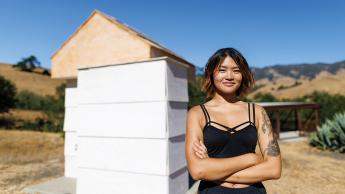 A young woman stands in front of a shed-like structure amid rolling hills