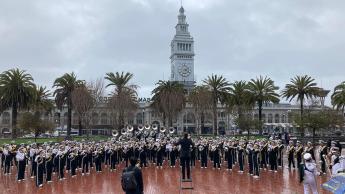 The Mustang Band performs in San Francisco's Embarcadero Plaza in front of the iconic Ferry Building on an overcast day.