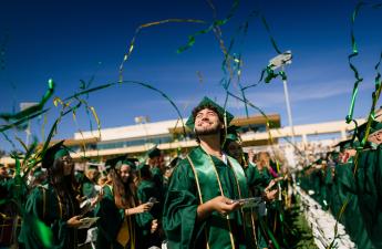 A student in green and gold graduation regalia smiles into the sky as green and gold streamers sail overhead.