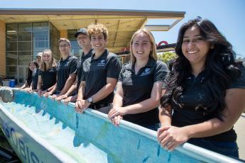 Seven smiling students line up for a photo alongside a light blue-tinted canoe