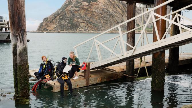 Three students putting on diving gear on a dock in Morro Bay.