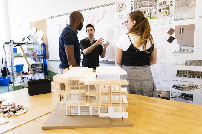 Two students talk with the professor against a display of building materials and behind an architectural model of a building. 